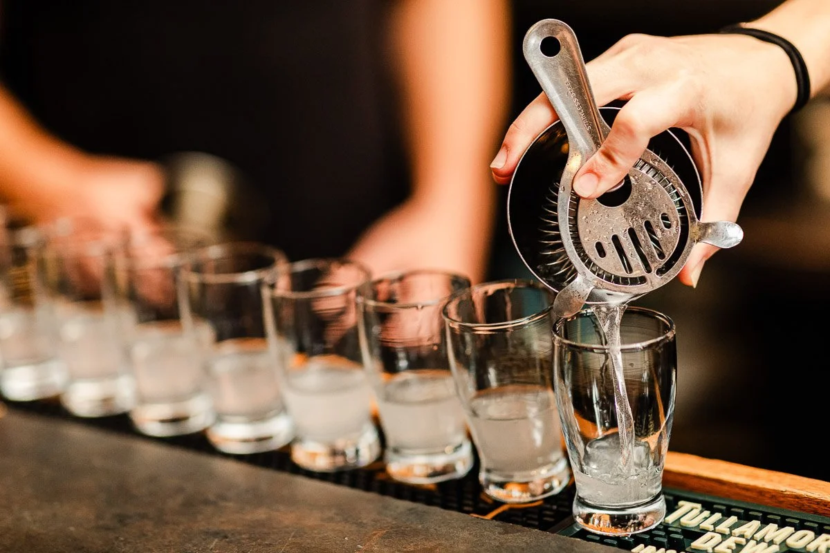 Hands pouring a clear liquid through a strainer into a row of shot glasses on a bar. The setting is lively and social, suggesting a bar scene.