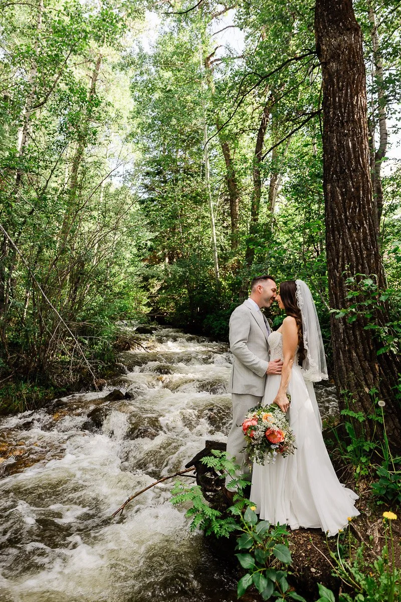A couple in wedding attire stands by a forest stream, surrounded by lush green trees. The bride holds a bouquet, and they lovingly gaze at each other captured by Beaver Creek wedding photographer tomKphoto
