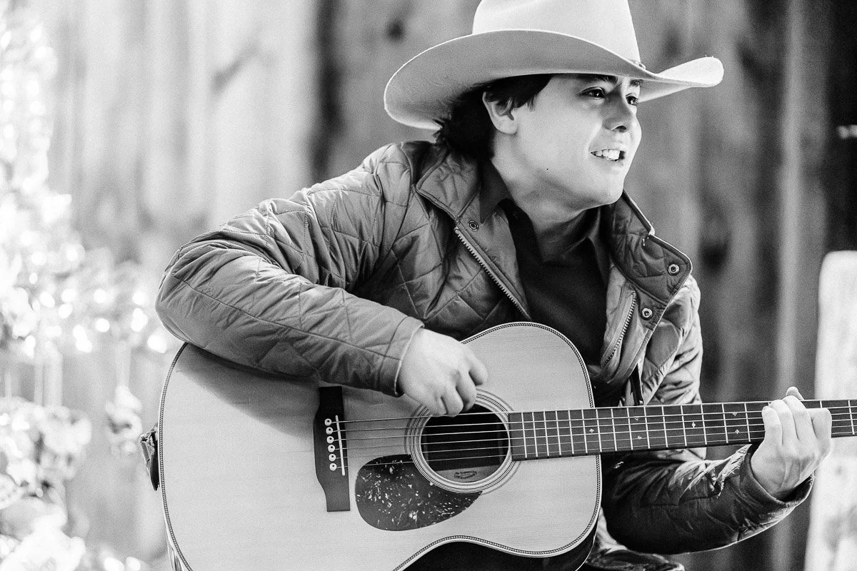 Black and white image of a person in a cowboy hat playing an acoustic guitar. They wear a quilted jacket and smile, creating a joyful, relaxed mood.