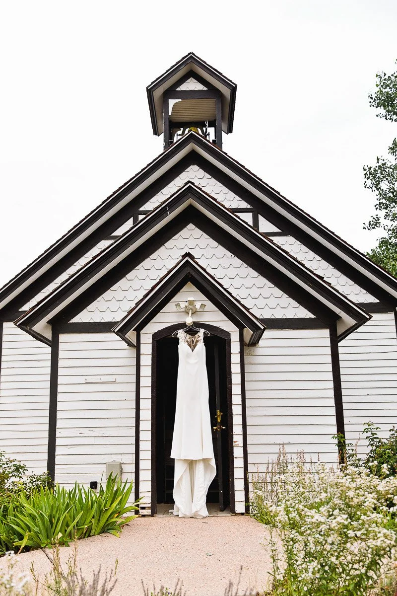 A white wedding dress hangs elegantly over the entrance of a quaint one-room schoolhouse with wooden siding and a small bell tower, surrounded by lush greenery.