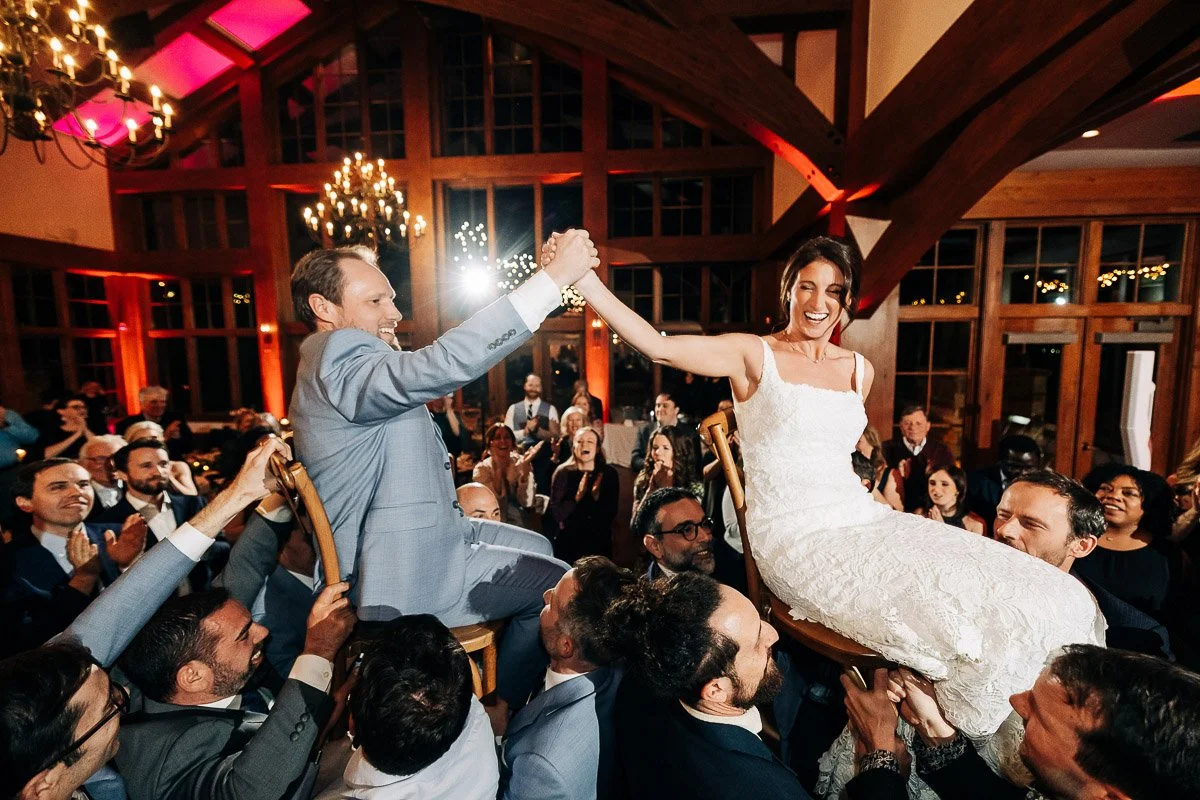 A joyful wedding scene with a couple lifted on chairs during the Hora dance. Guests surround them clapping, under chandeliers in a warmly lit hall.