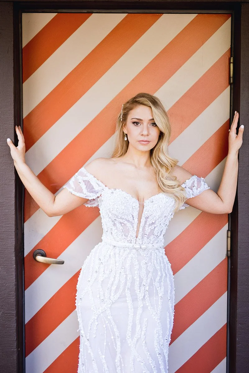 A woman in an off-shoulder white lace gown stands confidently in front of a door with diagonal orange stripes, conveying elegance and poise.