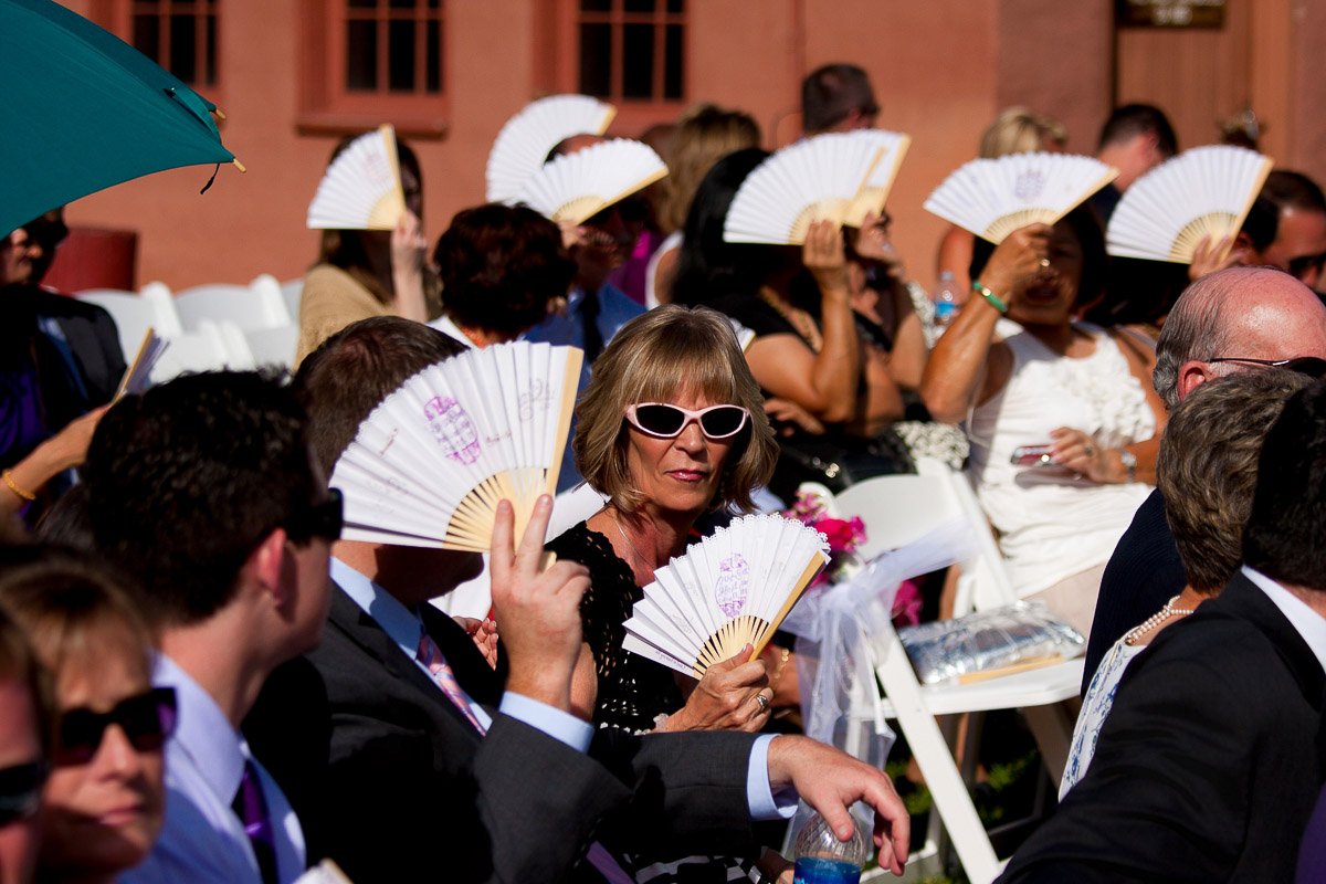 A group of people at an outdoor event, dressed in formal attire, sit in rows of white chairs holding decorative fans to shield from the sun.