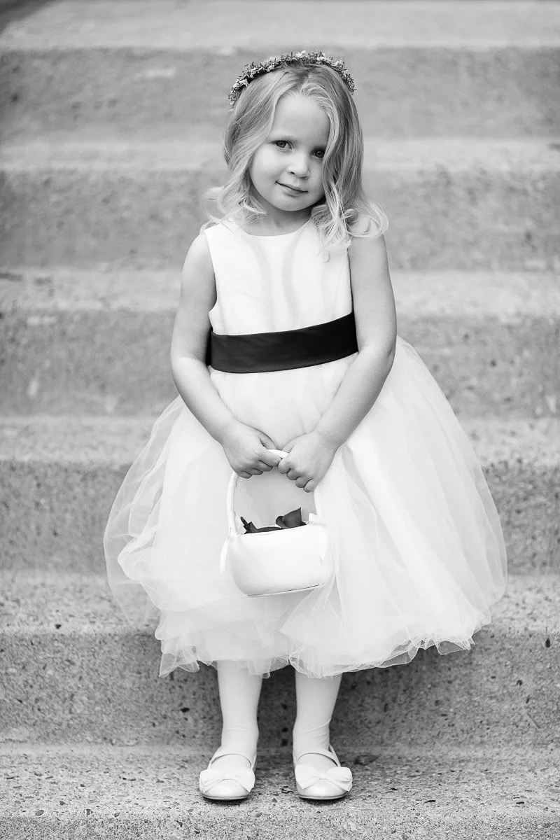 A young girl in a white dress with a black sash stands on steps, holding a small basket. Her gentle smile and floral headband convey an innocent charm.