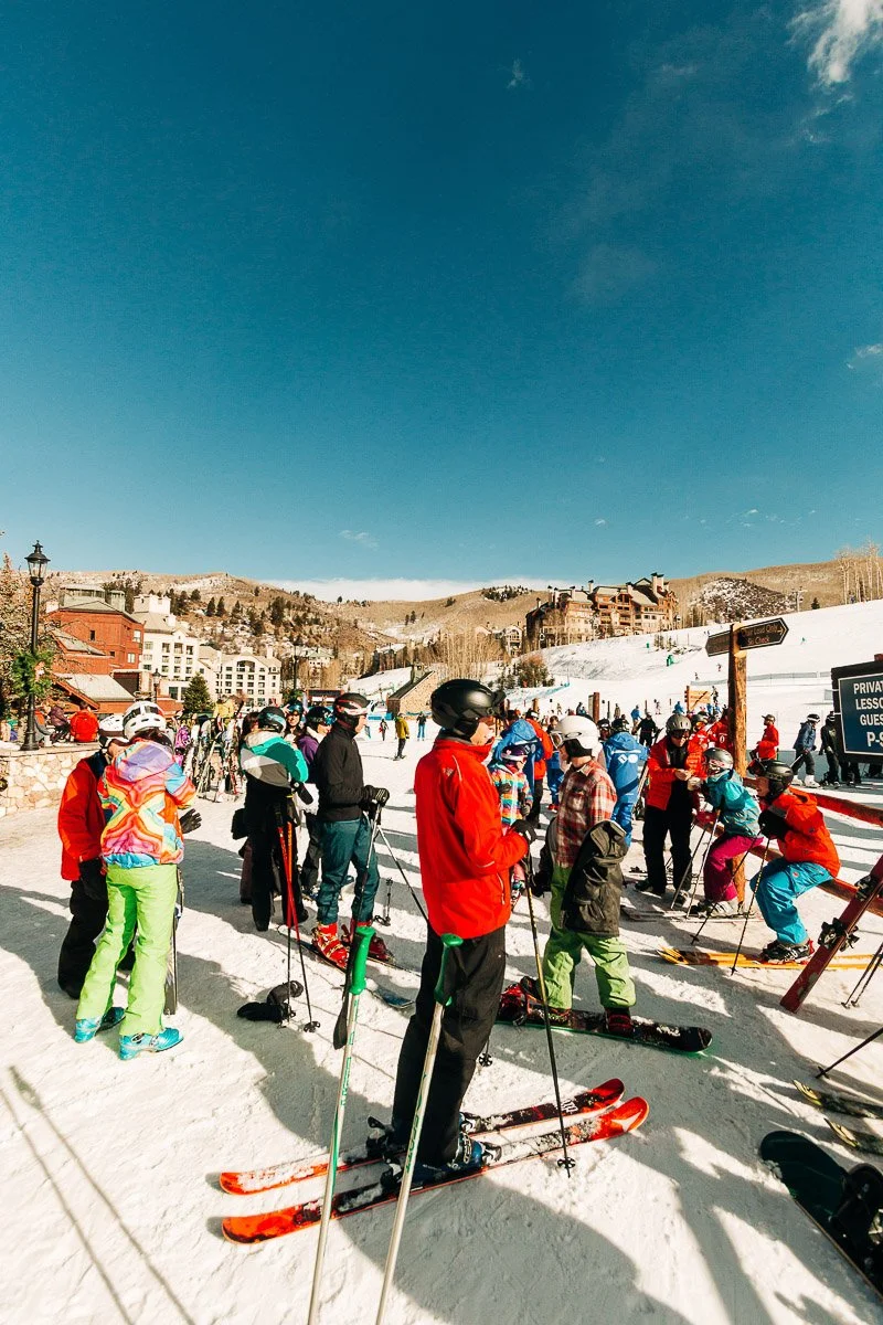 A lively ski resort scene with people in colorful winter gear and skis, gathered on a snowy slope under a clear blue sky, conveying excitement and activity.