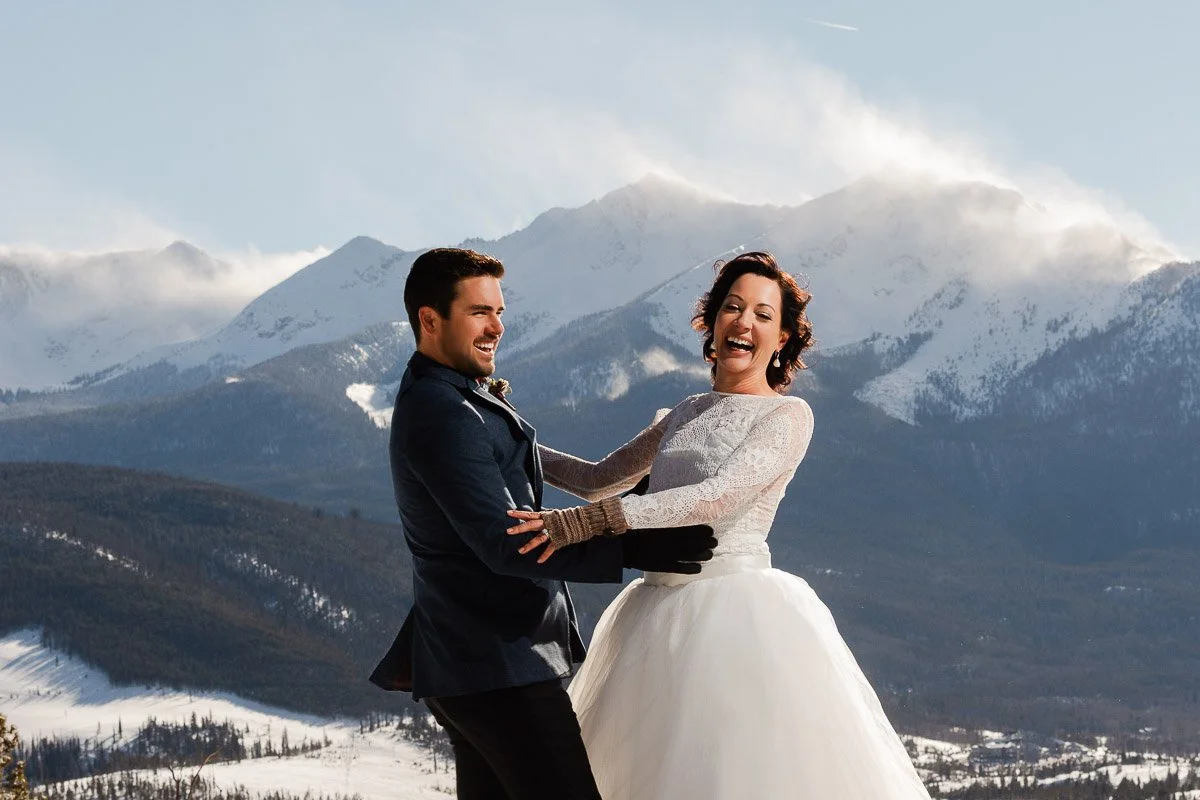 A joyful bride and groom in elegant attire laugh together against a stunning mountain backdrop with snow-dusted peaks and a clear blue sky captured by Colorado wedding photographer tomKphoto