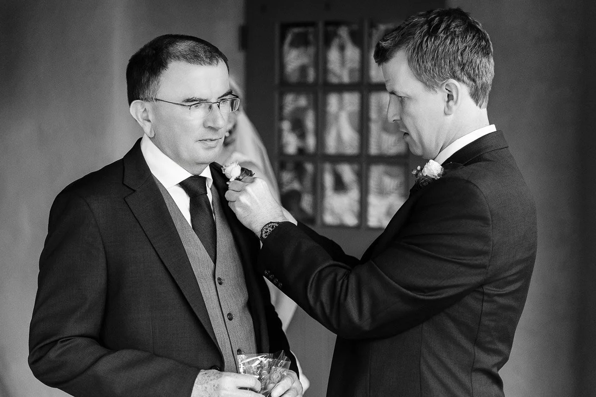 Two men in suits are shown in a warm, candid moment. One adjusts a boutonnière on the other's lapel. The setting conveys a formal, intimate occasion.