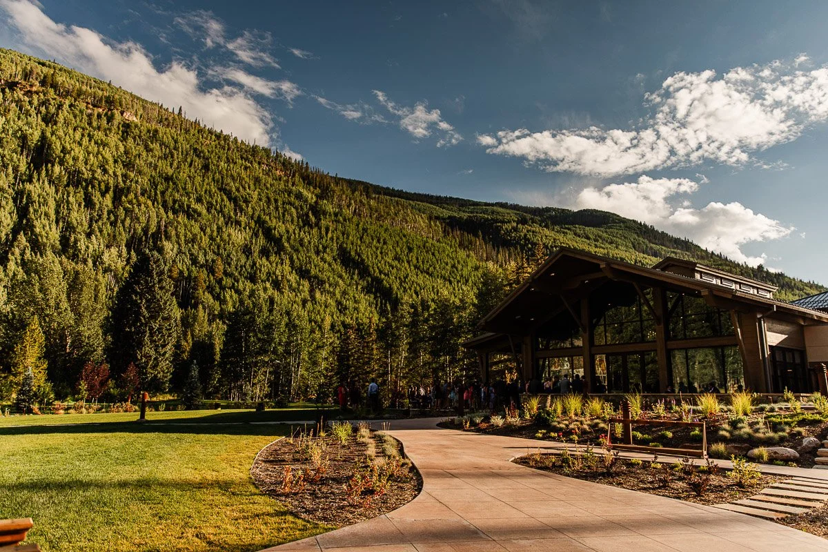 The Vail Golf Club with glass walls sits amidst lush green mountains and under a partly cloudy sky. A curved pathway leads to the entrance, surrounded by landscaped gardens.