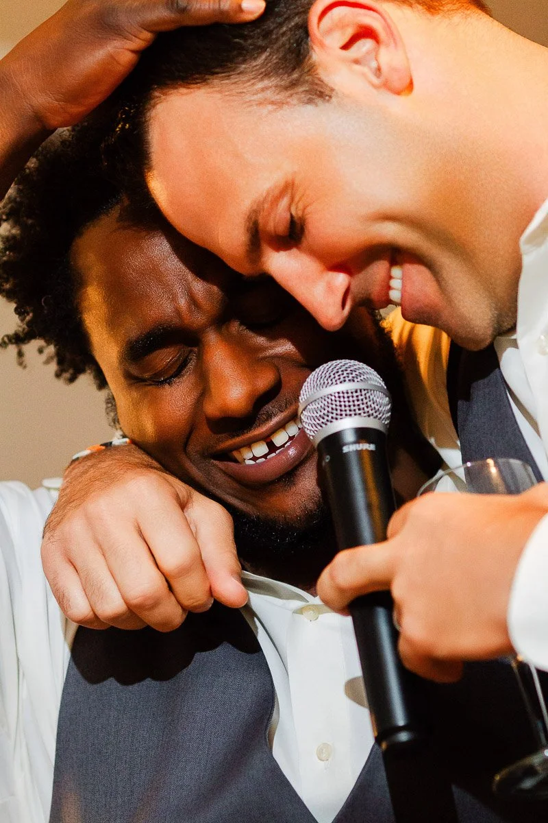 Two men in suits share an emotional embrace, one holding a microphone, both smiling warmly. The moment exudes joy and friendship.