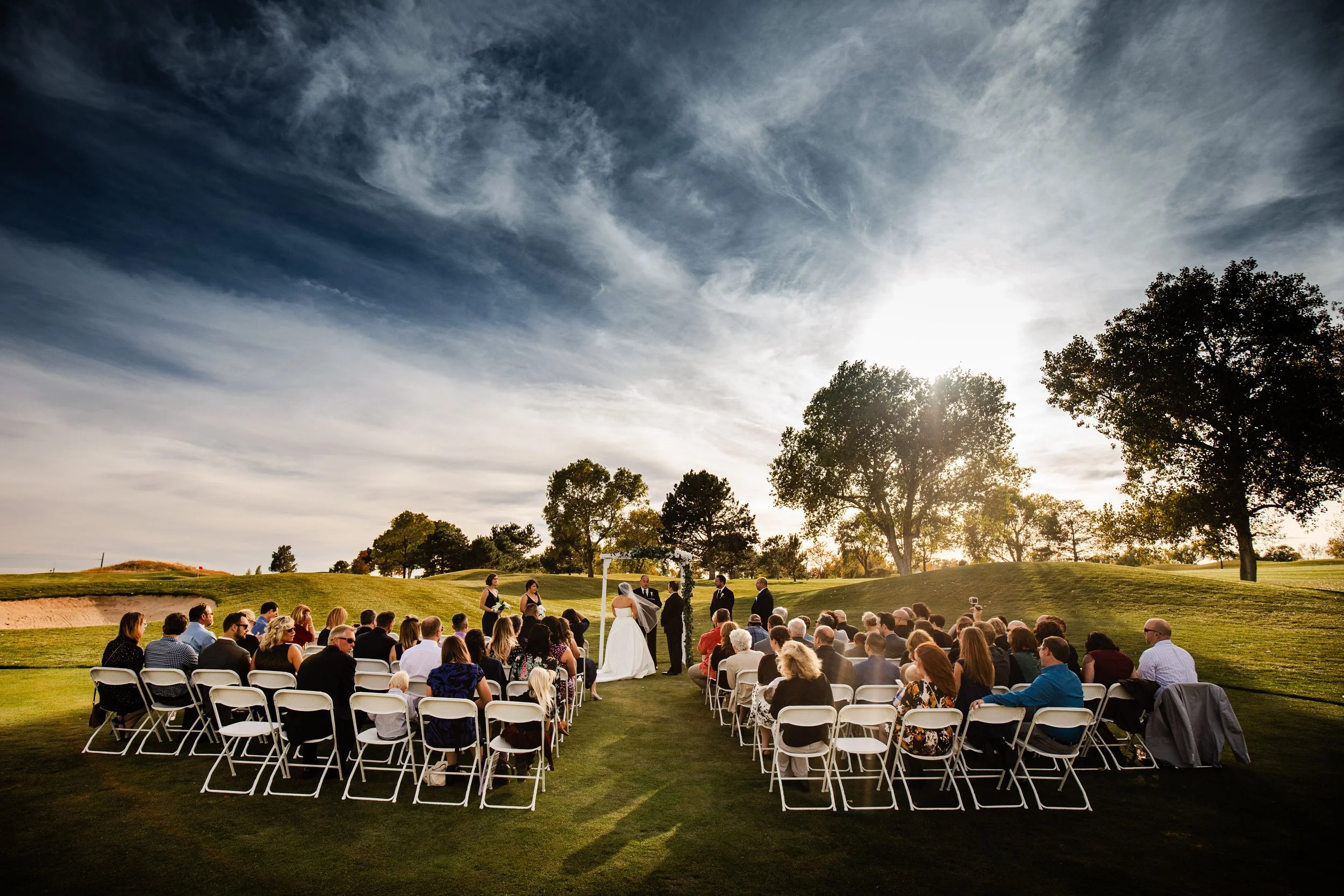 Outdoor Valley Country Club wedding ceremony on a grassy lawn. A couple stands under an arch, surrounded by guests seated on white folding chairs. Sunlight filters through trees.