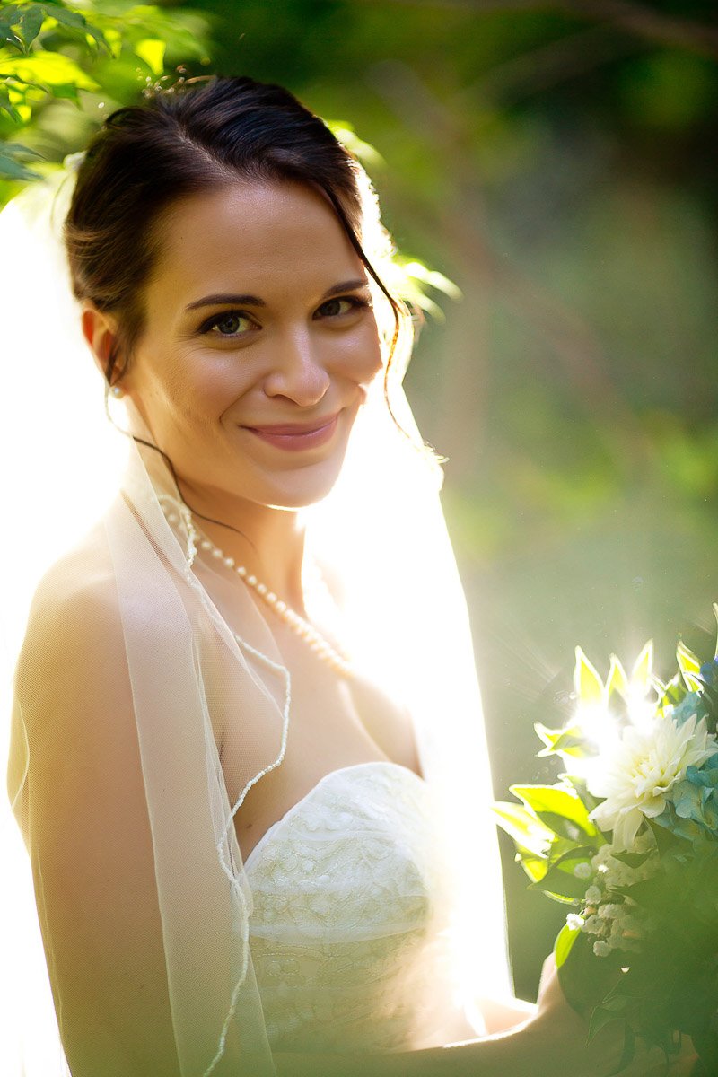 A radiant bride stands in sunlight, wearing a veil and strapless lace gown, holding a bouquet of white flowers. Her warm smile conveys joy and serenity.