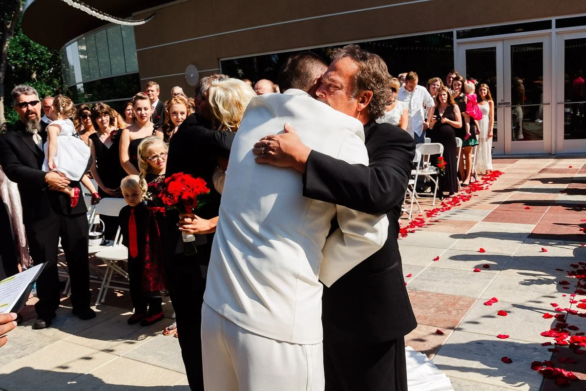 A groom in a white suit embraces a man in a black suit outdoors, surrounded by wedding guests. Rose petals are scattered along the path, creating a joyful atmosphere.