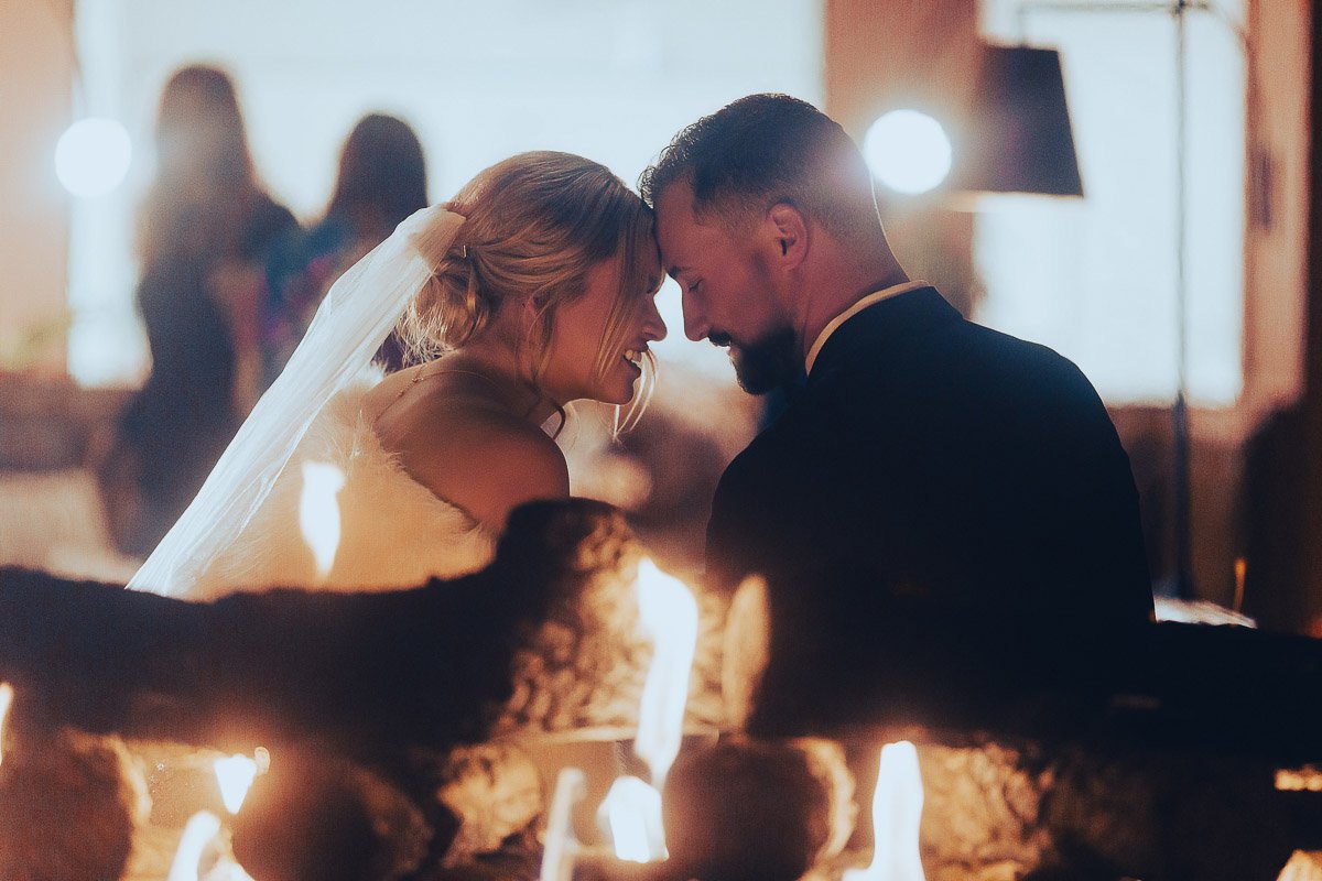 Bride and groom share a tender moment, foreheads touching, with warm firelight creating a romantic, intimate atmosphere.