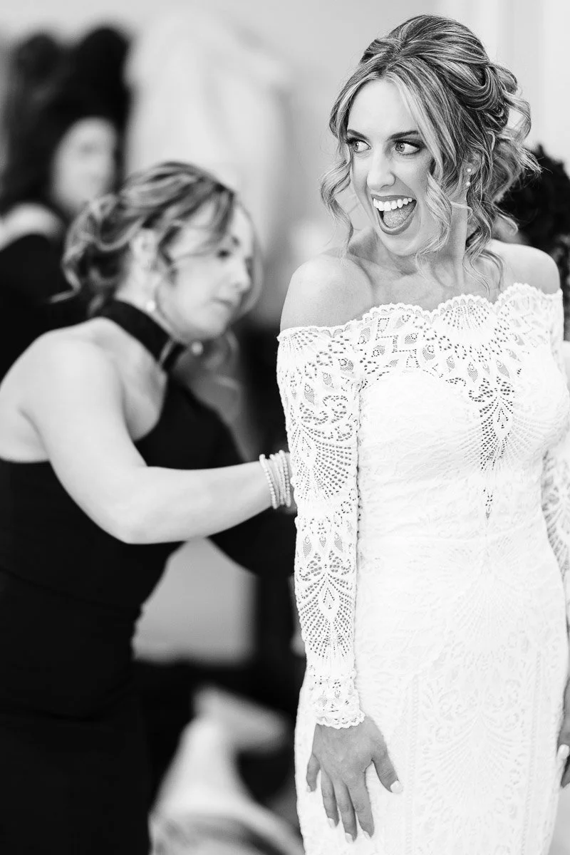 A joyful bride in an off-the-shoulder lace gown is being assisted by a woman in a black dress. The bride's expression is excited and happy.