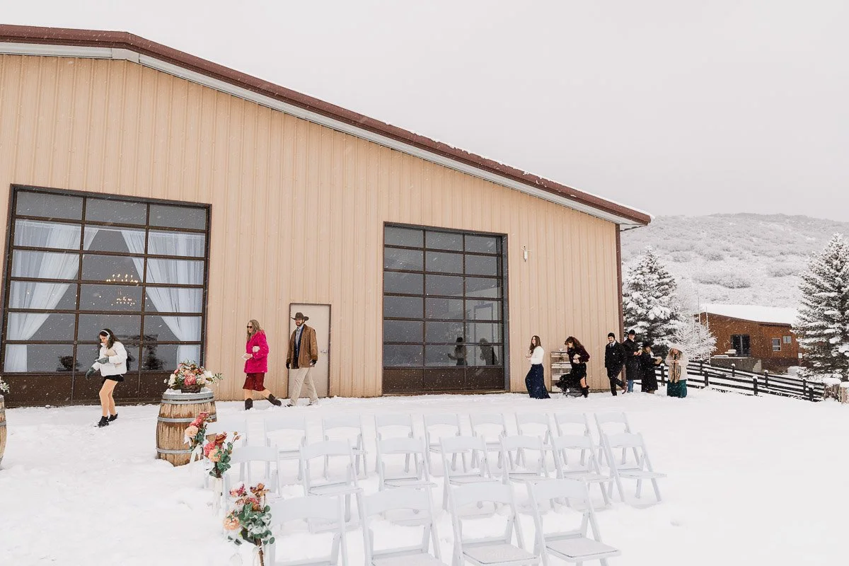 Guests in winter clothing walk outside a snowy beige barn venue with large windows. White chairs are arranged, and floral decorations sit on wooden barrels.