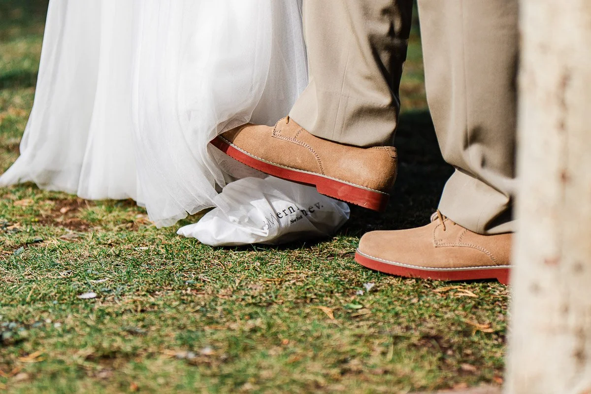 Groom in tan shoes steps on bride's white dress on leafy grass. Wedding scene exudes a playful, intimate tone. Formal attire highlights event.