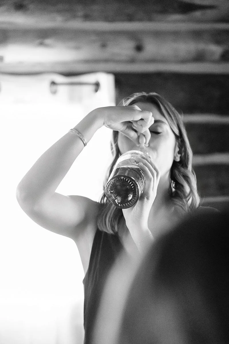 Black and white image of a person in casual attire taking a sip from a bottle indoors, with light streaming through a curtained window.