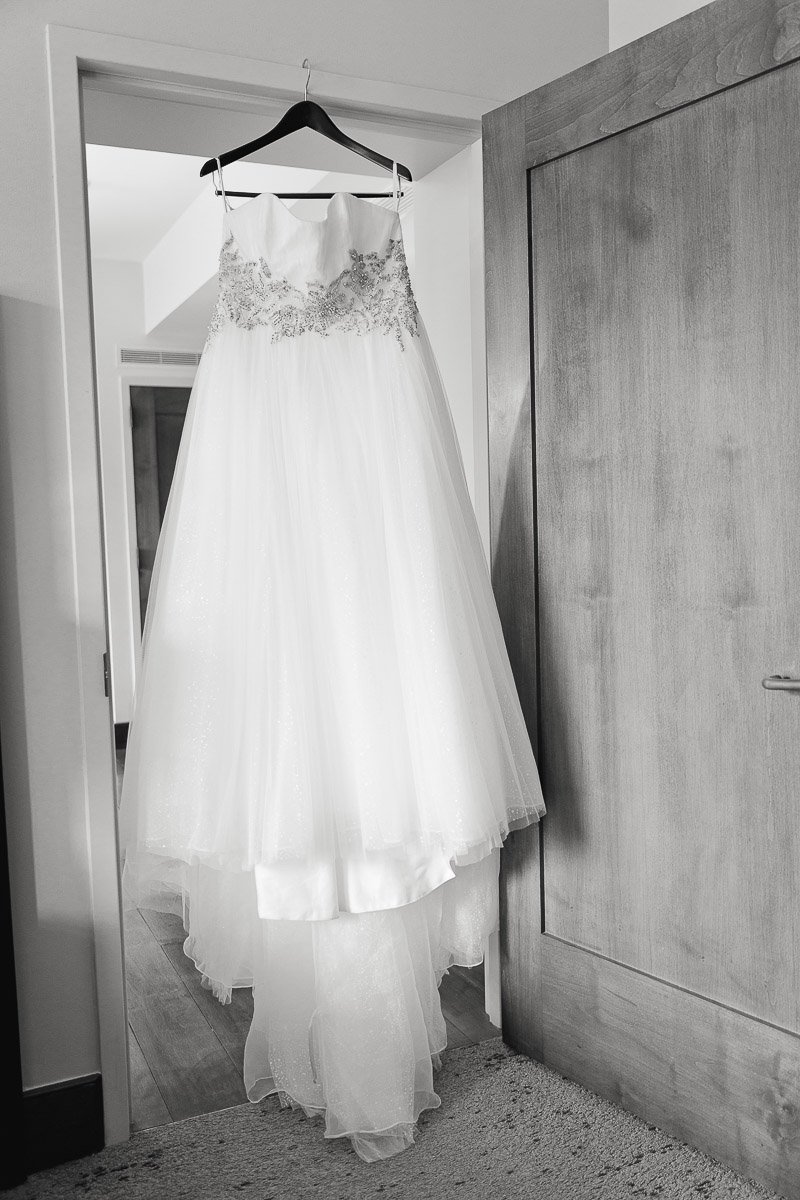 A white wedding dress with intricate lace detailing hangs on a door in a softly lit room. The elegant gown exudes a sense of romance and anticipation.