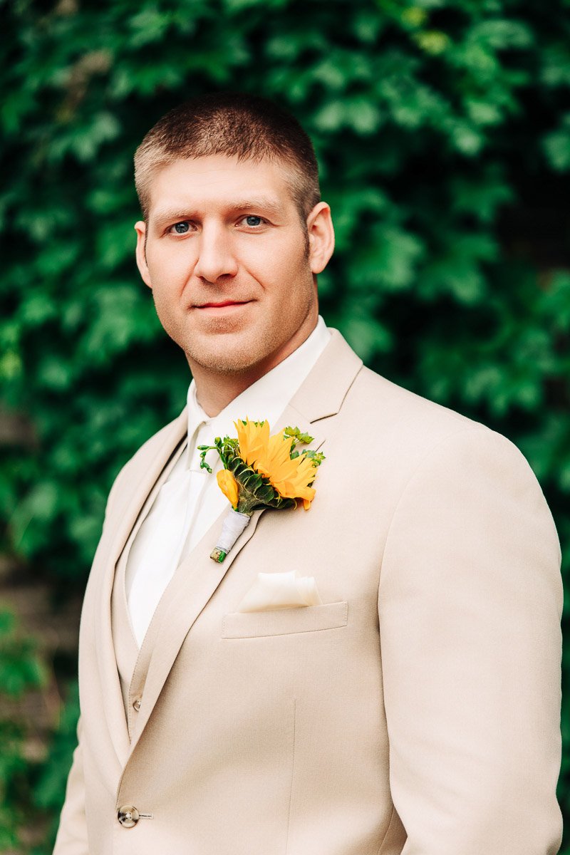 Man in a beige suit and white shirt stands confidently outdoors, with a yellow sunflower boutonnière. Lush green foliage forms the backdrop.