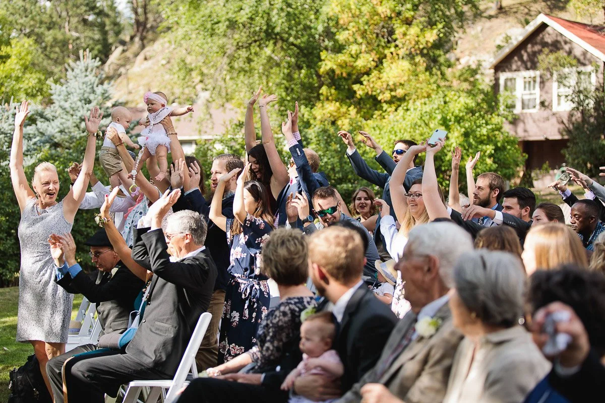 A joyful wedding scene with guests in outdoor seating, raising hands and cheering. Lush greenery surrounds the area; a house is visible in the background.