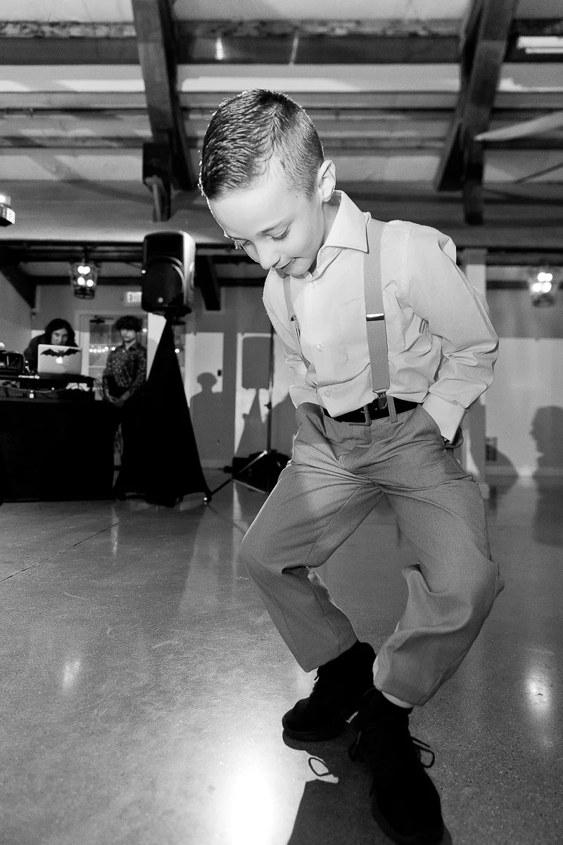 Young boy dancing energetically on a polished floor in a formal setting. Wearing a shirt, suspenders, and dress pants, he appears joyful and focused.