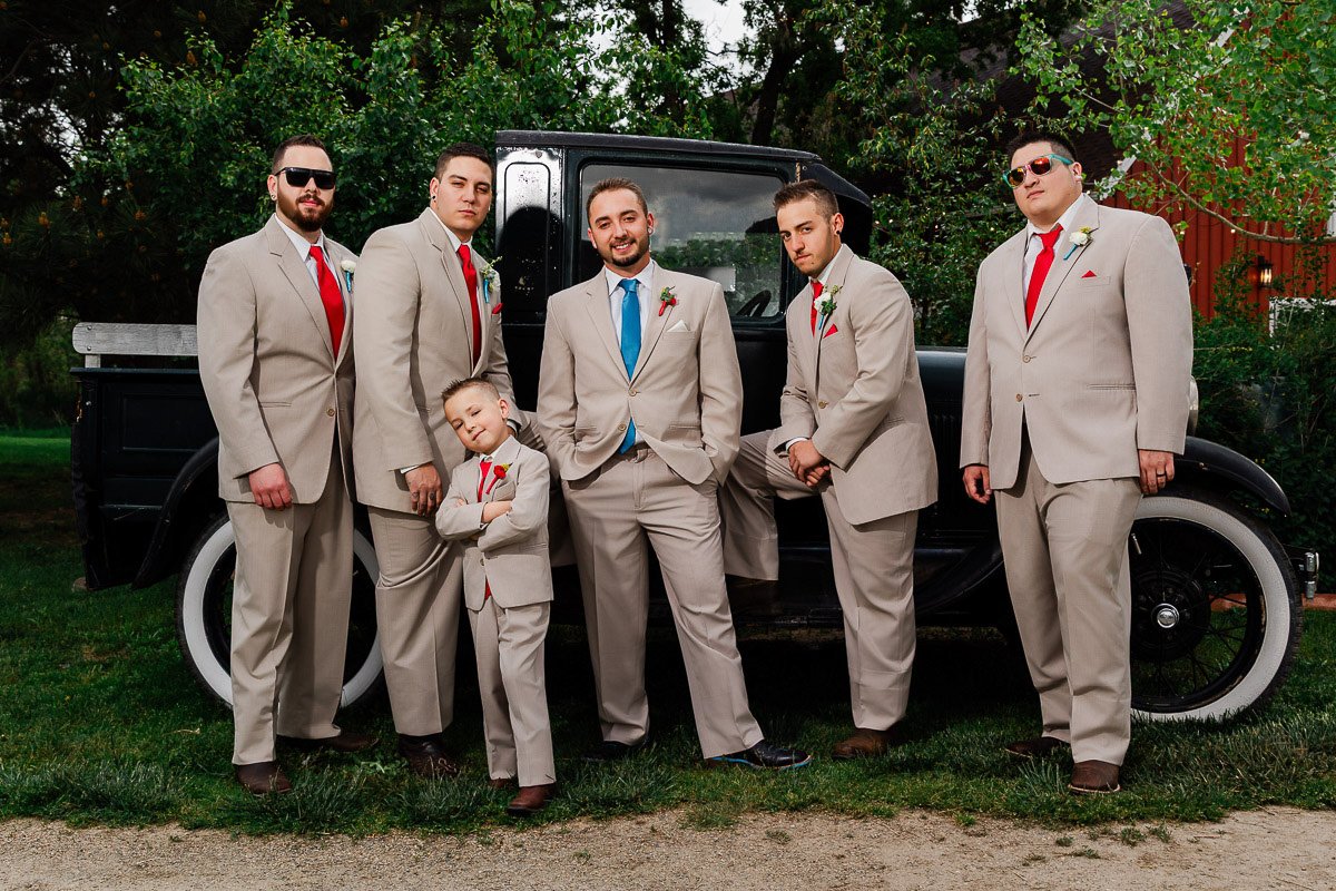 A group of five men and a boy, all in beige suits with red ties and boutonnieres, pose confidently in front of a vintage black car, exuding a celebratory and cheerful vibe.