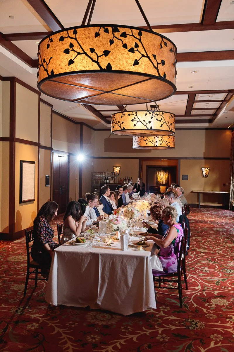 Dimly lit banquet hall with a long table set for dinner. Elegant chandeliers hang overhead, casting a warm glow. Guests seated, engaged in conversation.