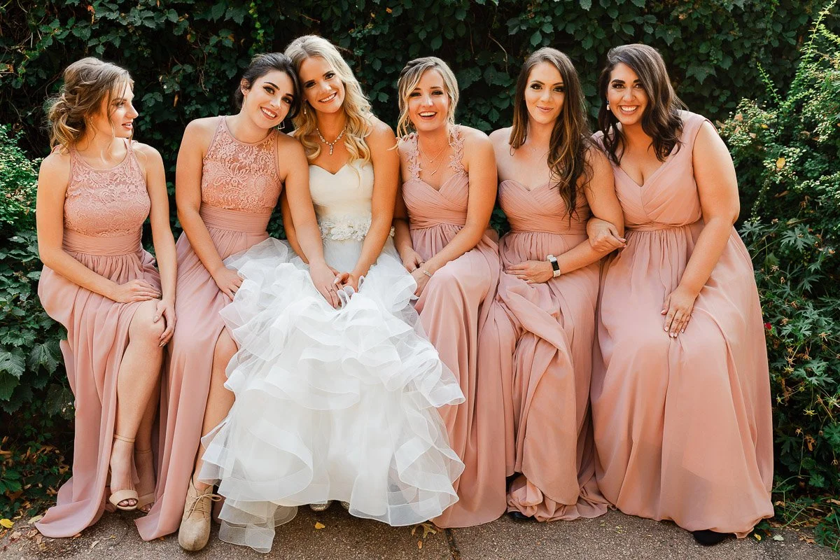 Seven women sit together, smiling brightly. The bride is in white, surrounded by bridesmaids in blush pink dresses against a leafy green backdrop during a The Lobby wedding reception in Denver