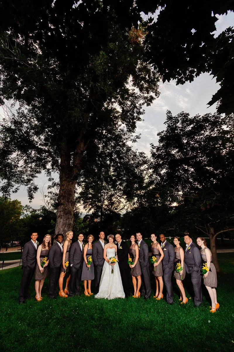 A bride in a white gown stands with bridesmaids in gray dresses and groomsmen in gray suits beneath towering trees, conveying a joyful, elegant outdoor wedding scene.