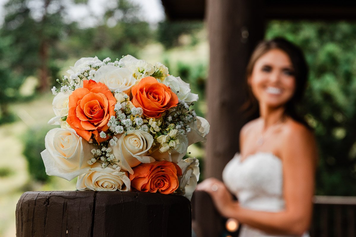 A vibrant bouquet with orange and white roses rests on a wooden rail. In the background, a blurred bride in a strapless gown smiles warmly.