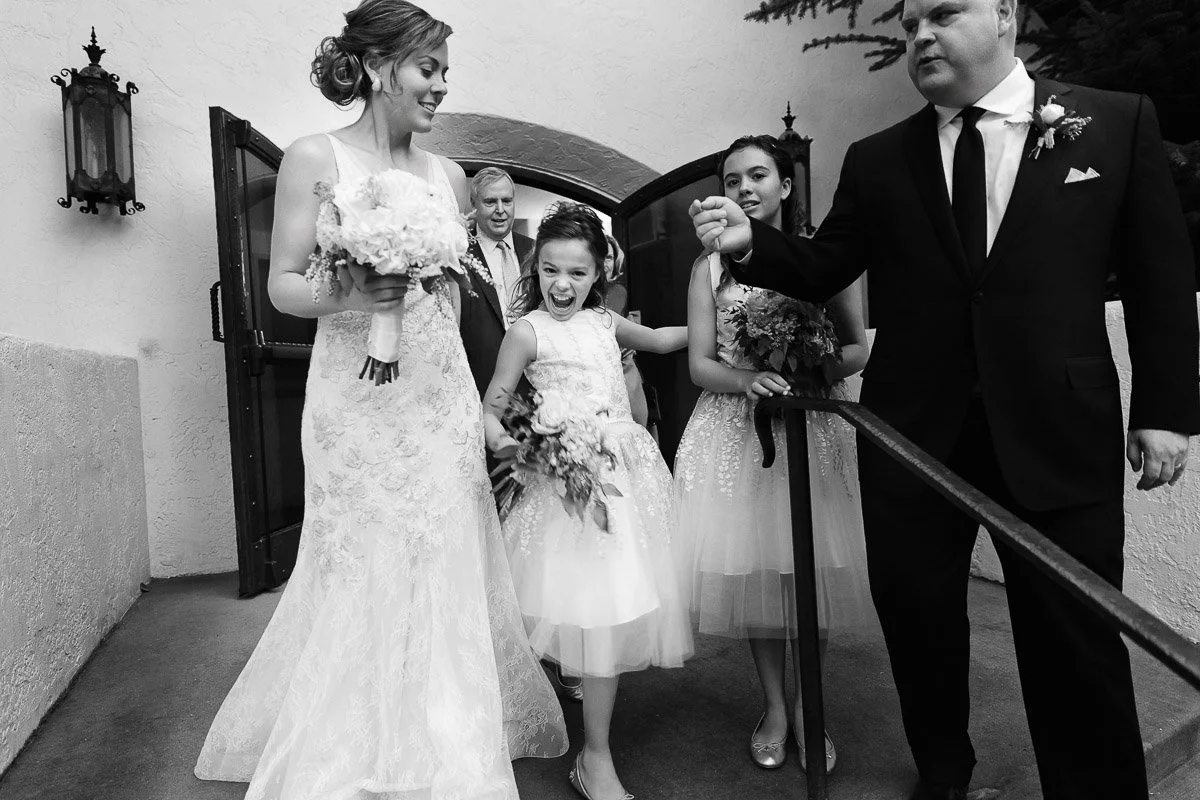 A bride in a lace gown exits a building, smiling beside a young girl with a joyful expression. A man in a suit and two bridesmaids accompany them. Classic, happy wedding moment.