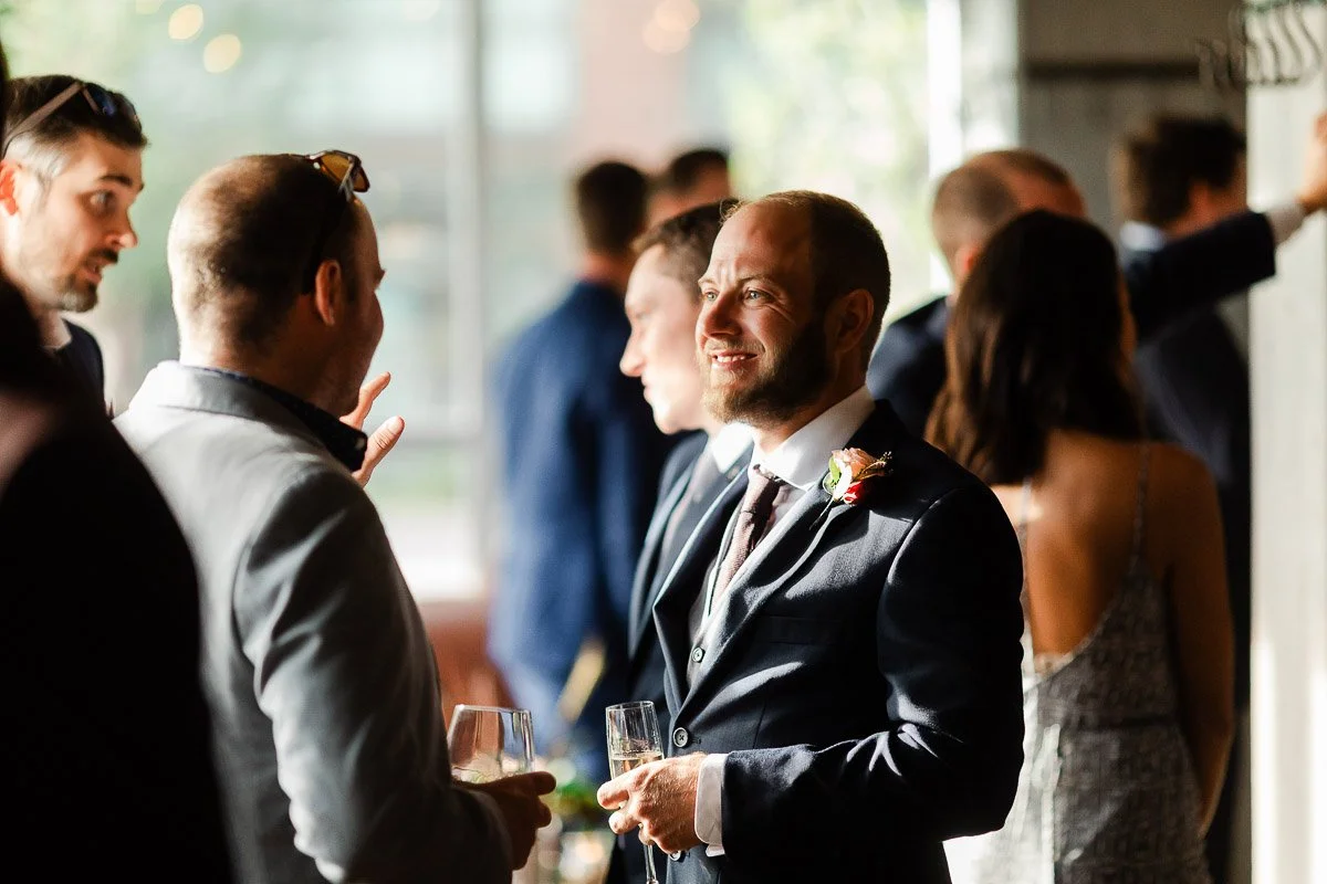 A man in a suit smiles while holding a drink at a lively, sunlit gathering. Others are engaged in conversation, creating a warm, social atmosphere.