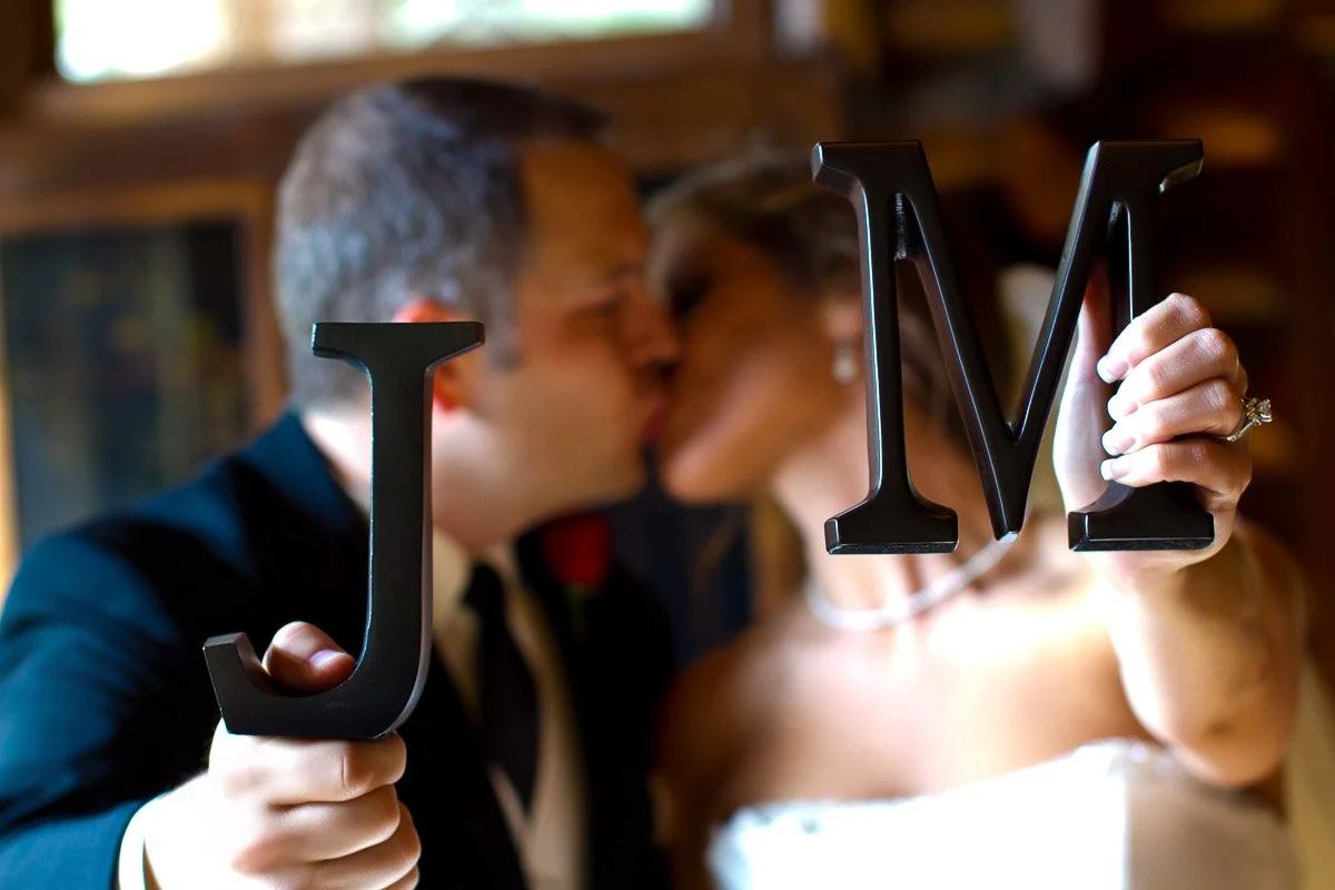 A bride and groom share a kiss while holding large decorative letters "J" and "M" in focus. The warm tones create a romantic, joyful atmosphere.