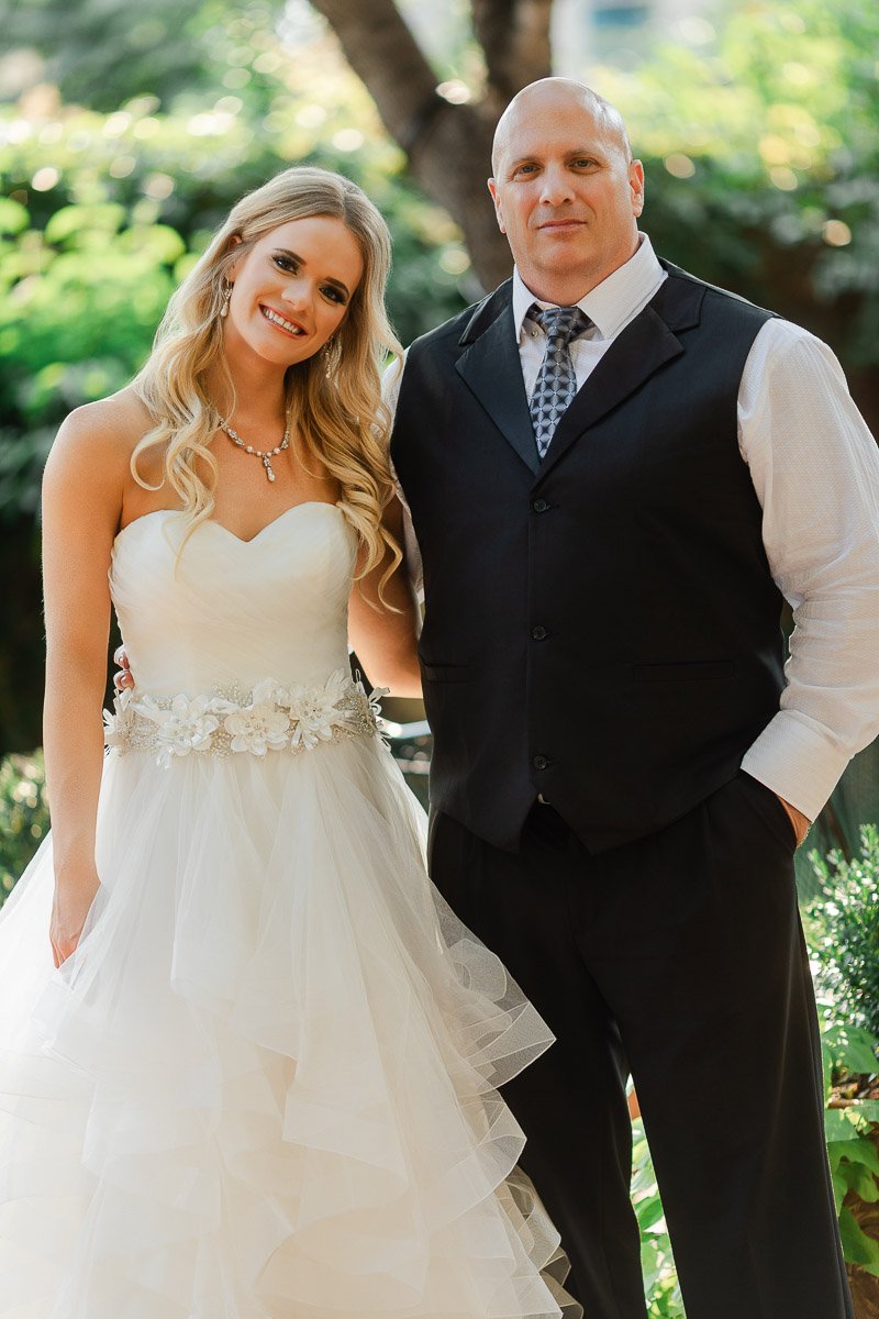 Bride in a strapless gown with floral detail stands beside a man in a black vest and tie. They smile warmly in a lush, garden setting.