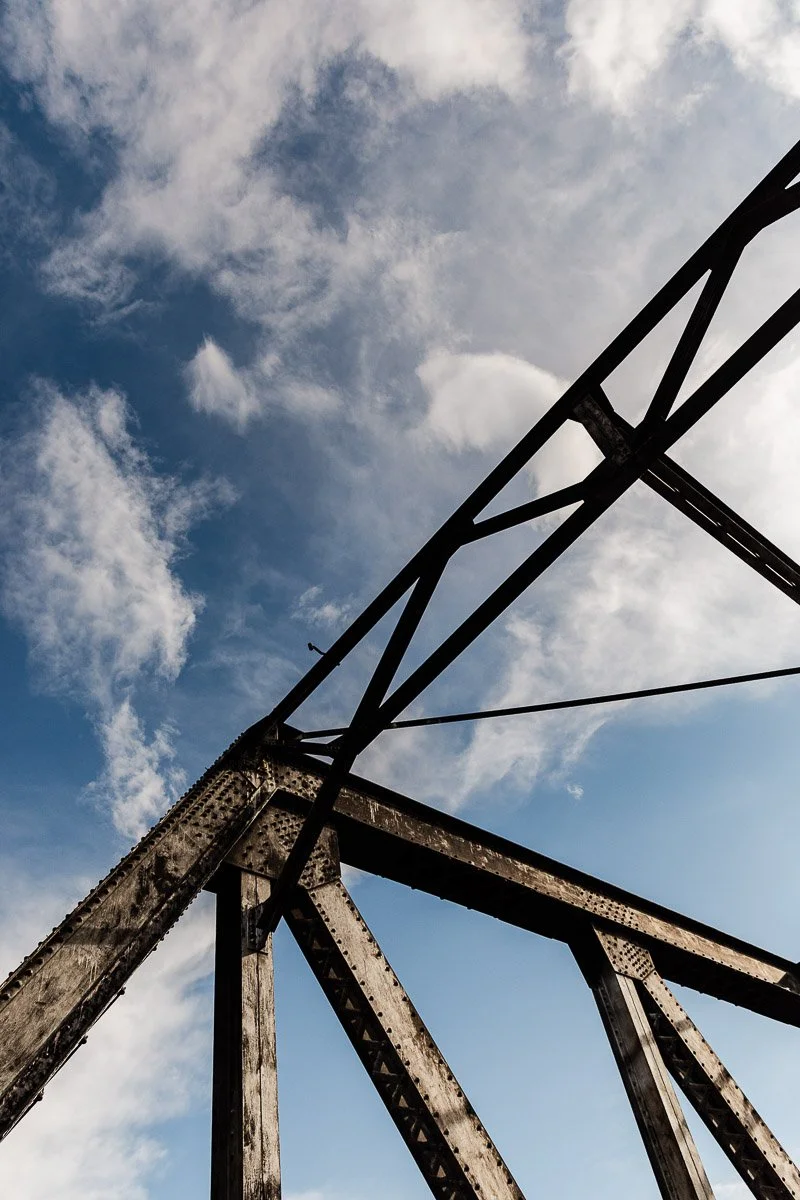 Rustic metal bridge supports against a vibrant blue sky with wispy clouds, creating a contrast between industrial structure and nature's expanse.