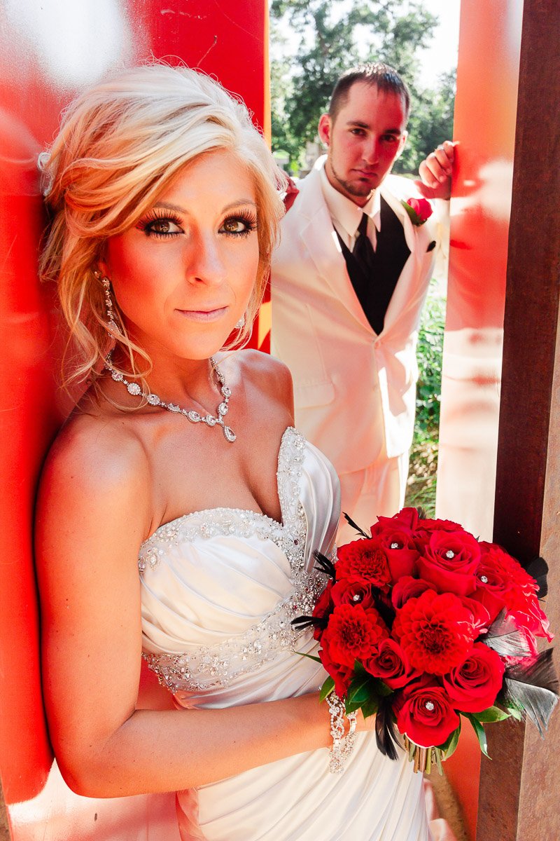 A bride in a white, jeweled dress holds a vibrant bouquet of red flowers. A groom in a white suit and red boutonniere stands behind her, closely positioned.