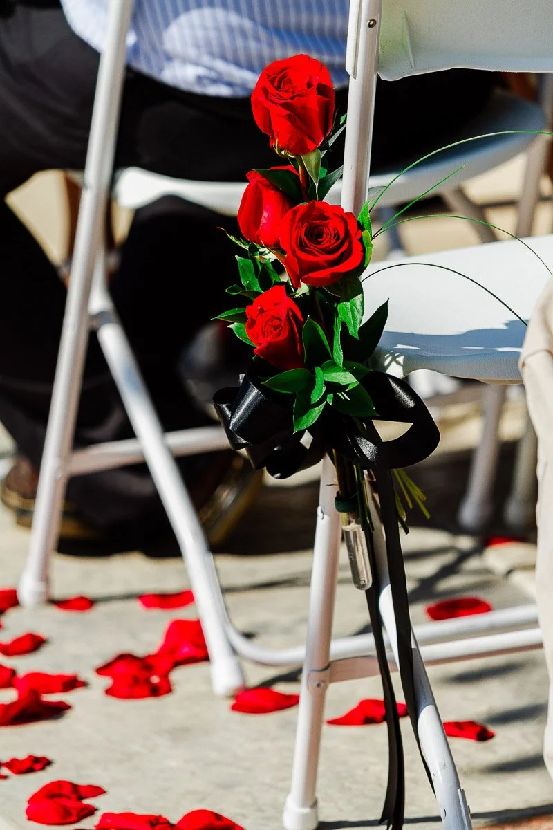 Close-up of three red roses tied with a black ribbon to a white folding chair at an outdoor event, with scattered rose petals on the ground.