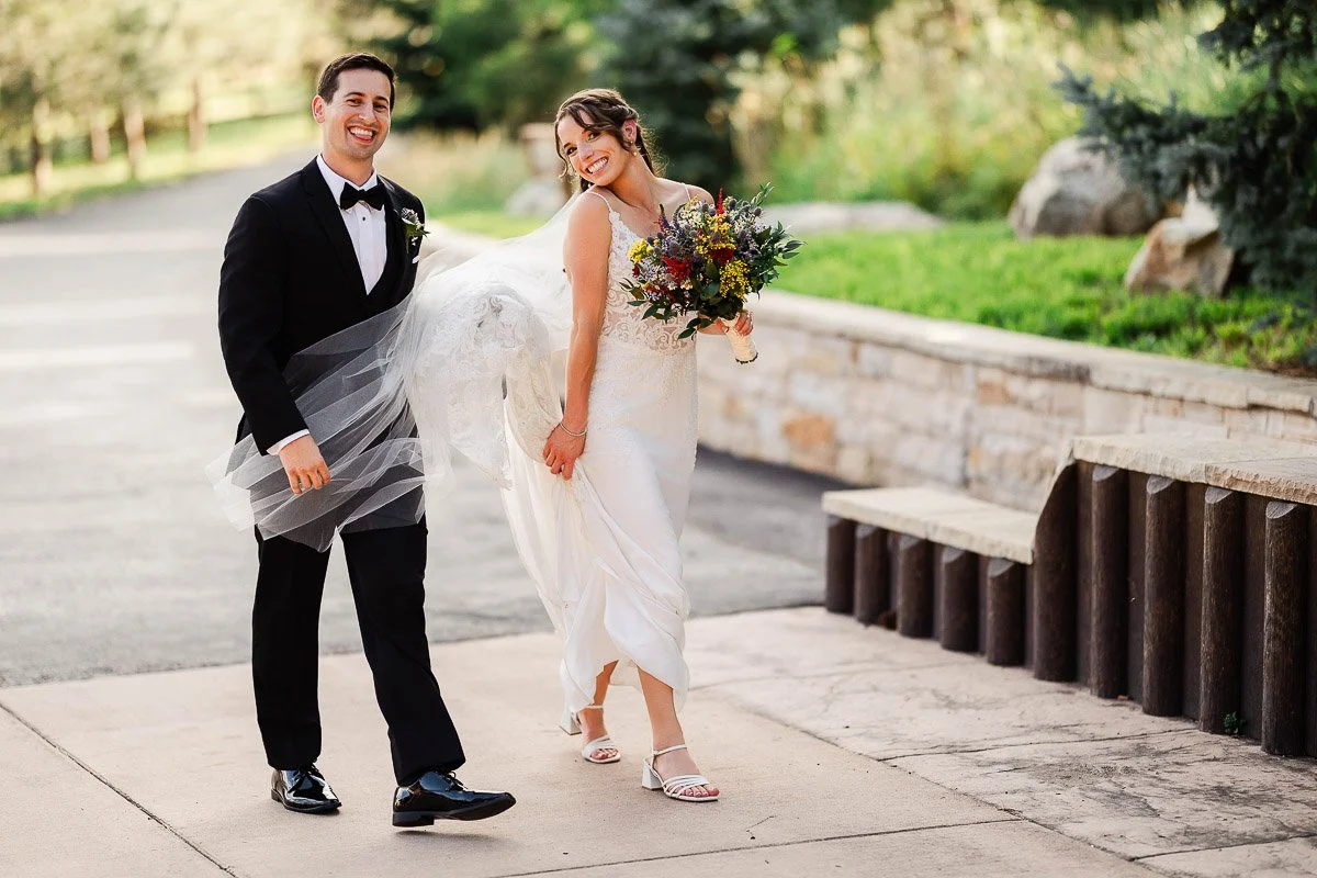 A joyful bride and groom walk outdoors. The bride in a white gown holds a colorful bouquet, while the groom in a black suit holds her dress.
