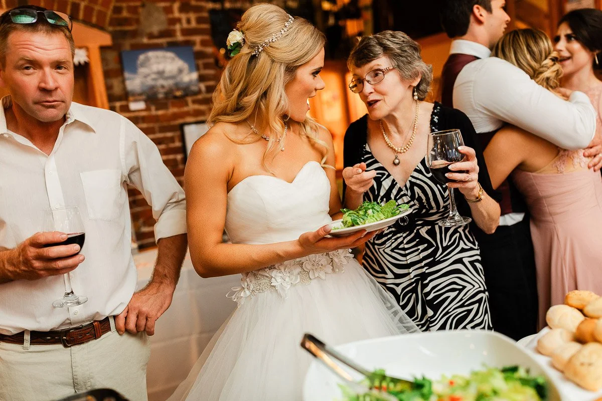 A bride in a strapless white dress and a guest in a zebra-patterned dress chat warmly, both holding wine glasses. Background displays a lively celebration.