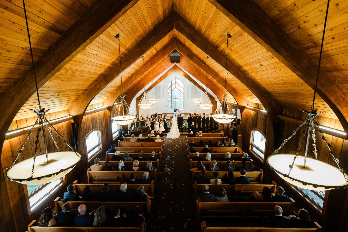 A bride and groom stand at the altar in a wooden church with vaulted ceilings and chandeliers. Guests seated in pews watch the ceremony.