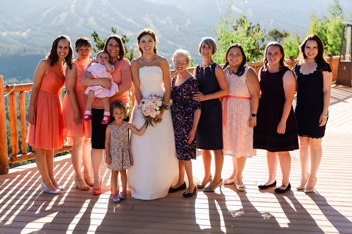A bride in a white gown is surrounded by nine smiling women on a sunlit wooden deck with a mountain view. They're dressed in colorful, semi-formal attire.