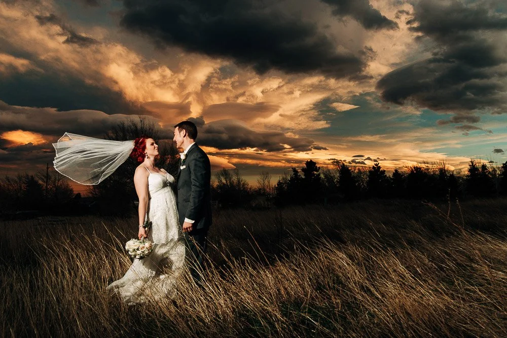 A bride and groom stand in a golden field at sunset, with dramatic clouds in the sky. Her veil flows in the wind, creating a romantic, intimate scene.