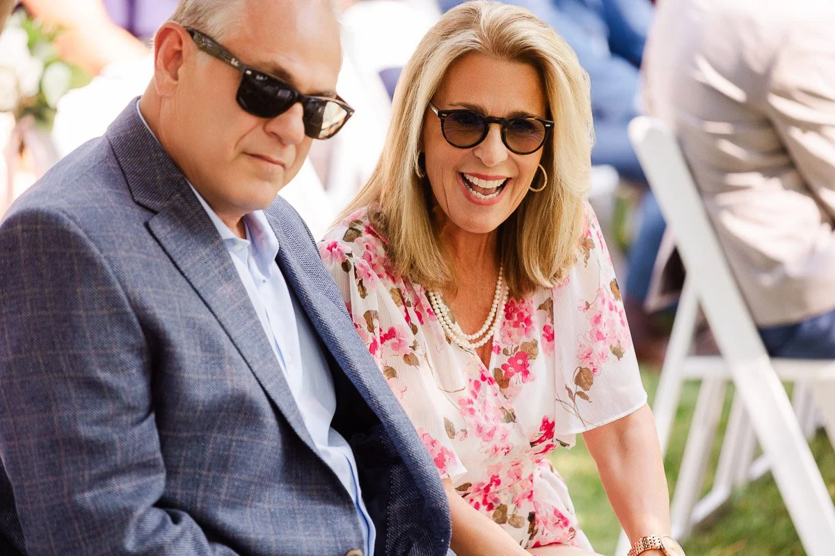 A smiling woman in a floral dress and sunglasses sits next to a man in a blue suit and sunglasses. They are outdoors on white folding chairs.