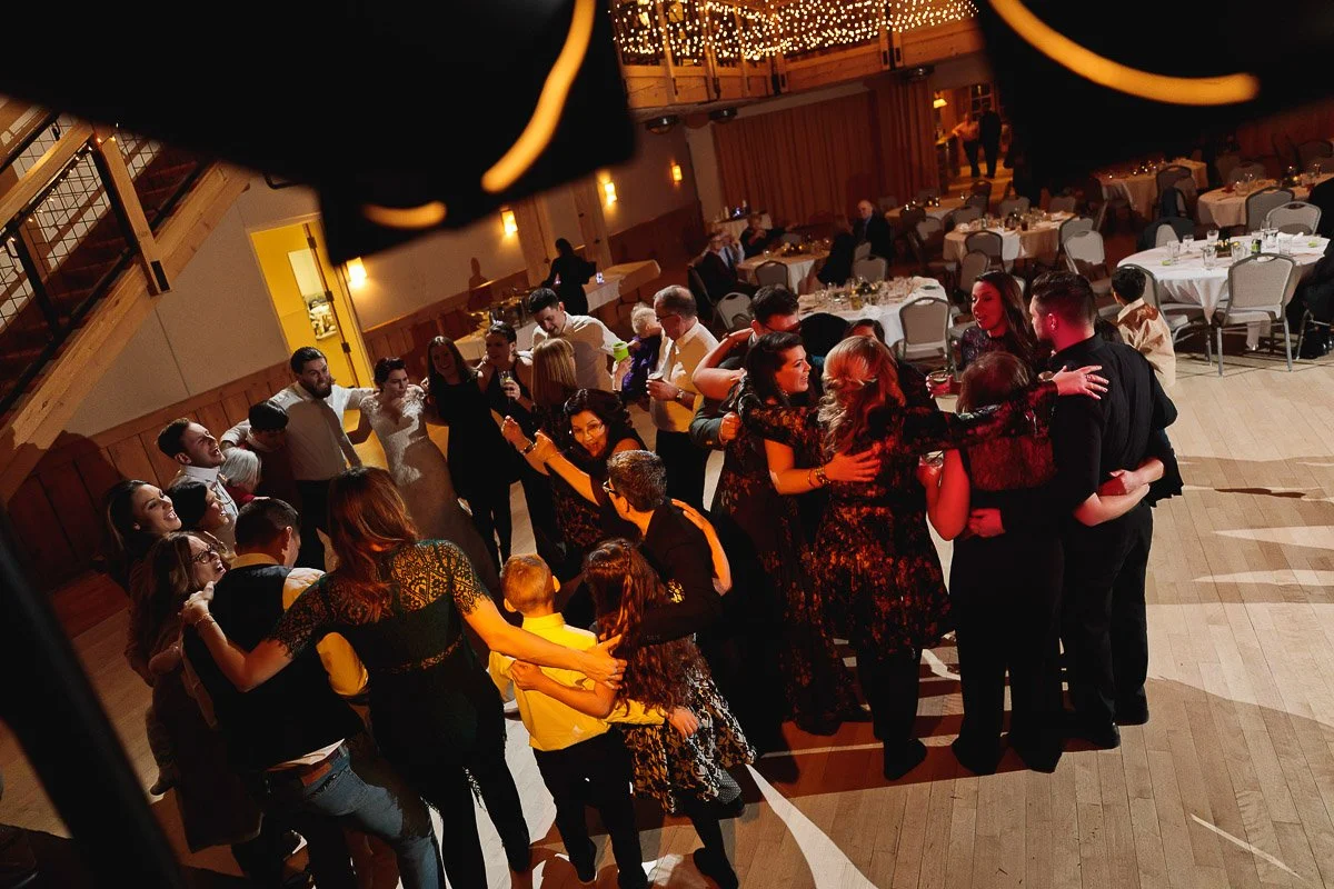 A joyful group of people stands arm-in-arm in a warmly lit banquet hall. String lights and decorated tables fill the spacious wooden venue.