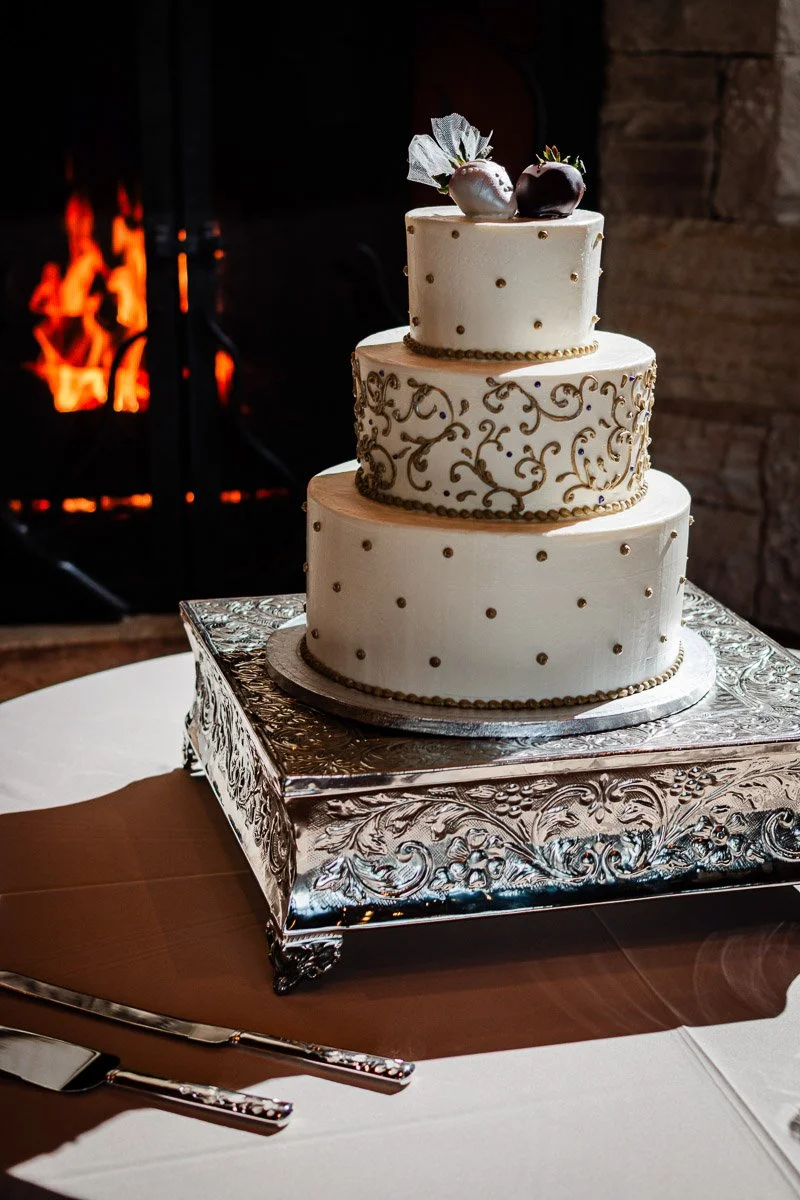 A three-tiered wedding cake with gold detailing and chocolate-covered strawberries on top, set on an ornate silver stand, with a warm fireplace glow in the background.
