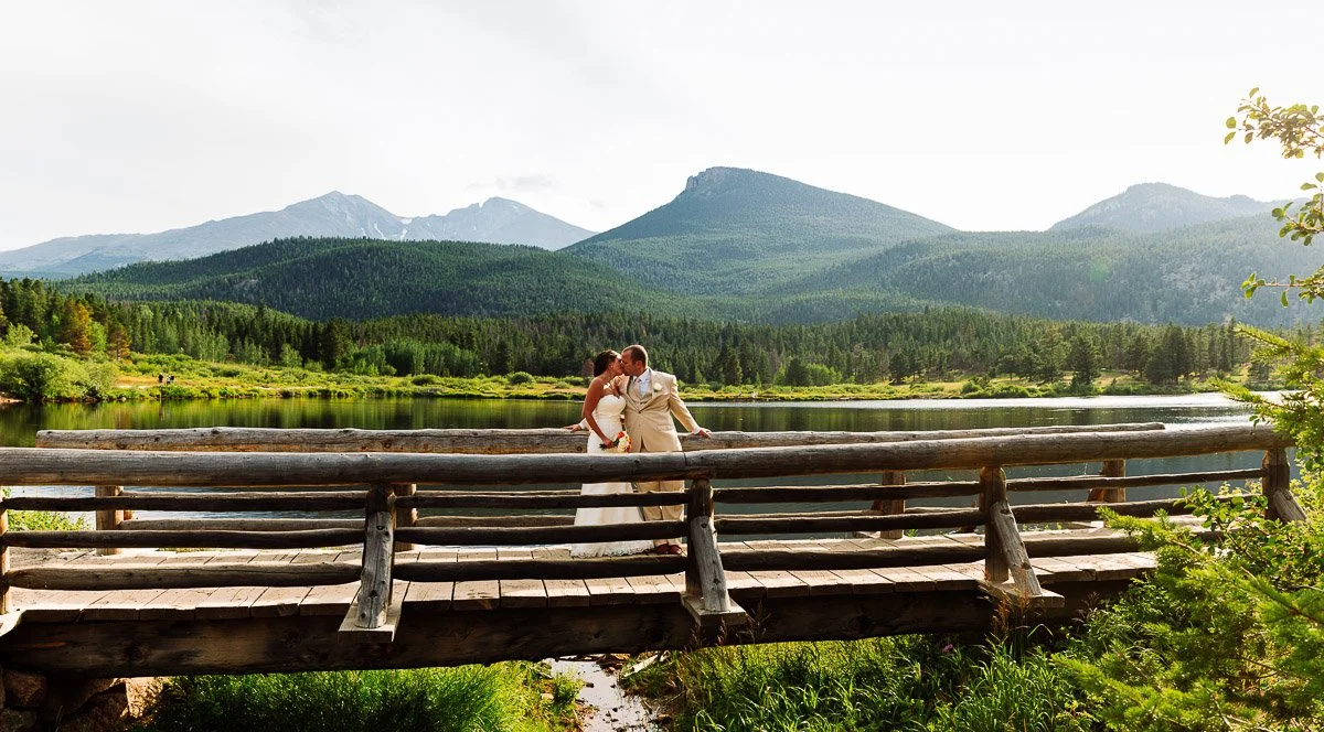 A couple in wedding attire shares a kiss on a rustic wooden bridge overlooking a serene Lily Lake, with lush forests and majestic mountains in the background.