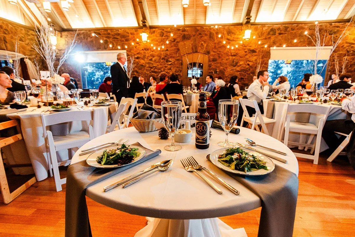 Elegant wedding reception scene with round tables, white chairs, and warm lighting. Tables are set with salads, drinks, and cutlery, creating a celebratory and inviting atmosphere.
