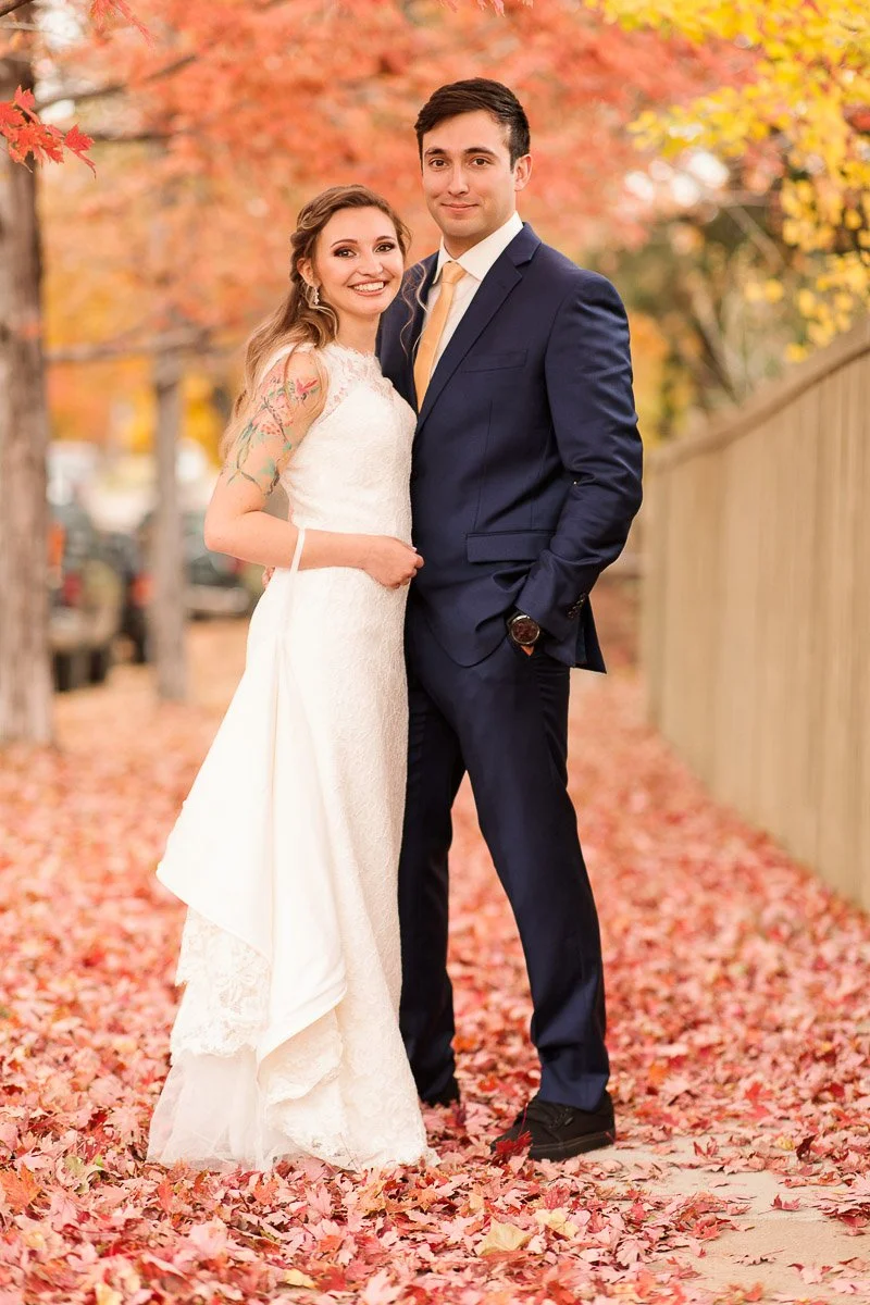 A couple stands on a path covered with red and yellow leaves. The smiling bride wears a white dress, and the groom wears a navy suit. The scene feels joyful and romantic.