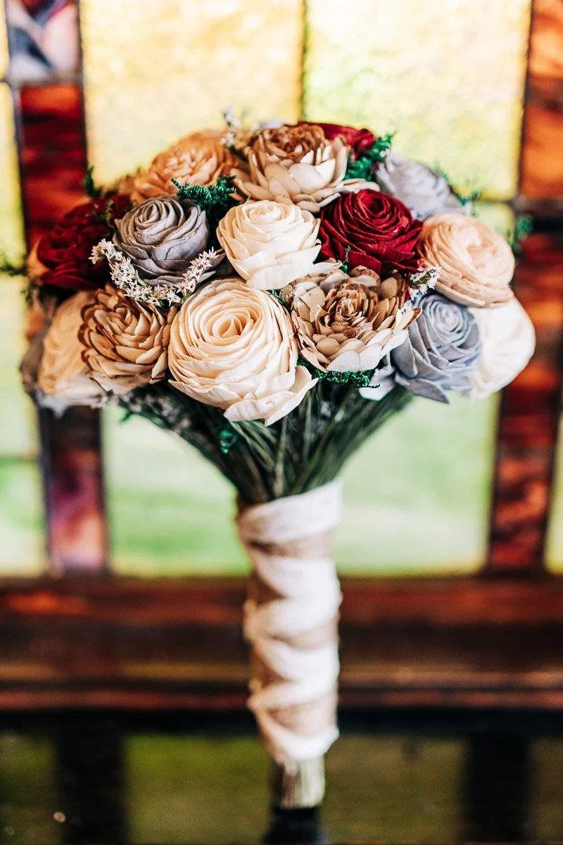 A bouquet of cream, red, and gray roses with green accents, wrapped in twine, against a blurred stained glass background, conveying elegance and warmth.