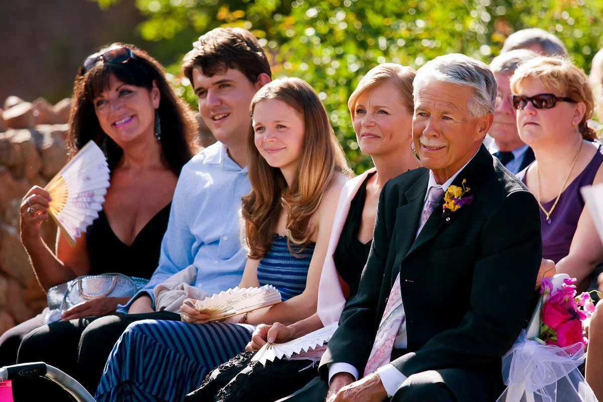 A group of smiling people, including a man in a suit with a purple boutonnière, sit outdoors at an event. They hold fans, enjoying the sunny weather.